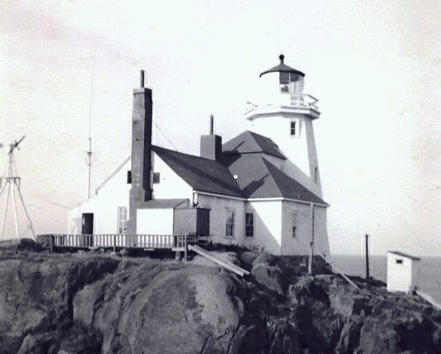 Black-and-white photograph of the Egg Island light station: a white tower with a
                    lantern gallery rising behind a shingled keeper's house, both set on bare rock
                    above a plank walkway and railing.
