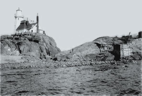 Black-and-white photograph of Egg Island from the water: the lighthouse and its
                    house standing on a low rock headland, with a timber landing and derrick built
                    against the rocks to the right and a shingle beach between them.