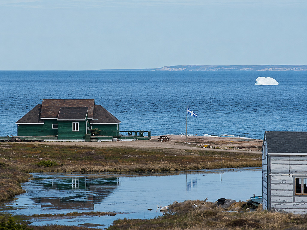 A green clapboard house on the shore at Blanc-Sablon, the Québec flag flying
                  beside it, with an iceberg drifting in the Strait of Belle Isle beyond.