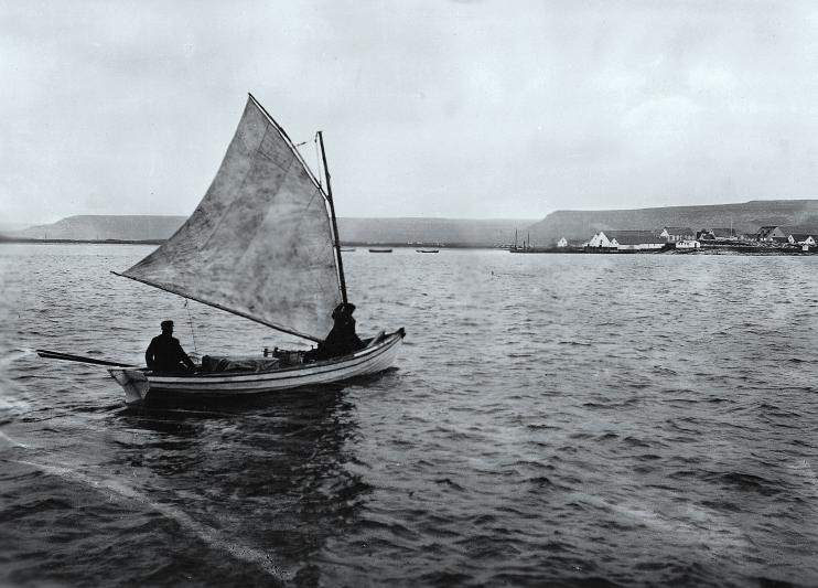 Two men sail a small open fishing boat under a patched sail, the village of
                  Blanc-Sablon visible on the far shore.