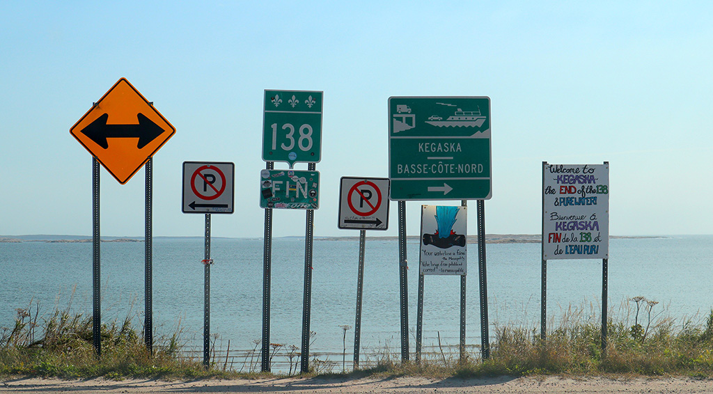 A cluster of tall roadside signs standing against open water at the end of a
                  gravel road: a Route 138 shield, a green panel reading Kegaska Basse-Côte-Nord,
                  and a hand-lettered board welcoming visitors to the end of the 138.