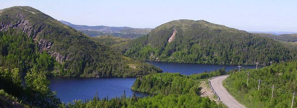 A gravel-shouldered road curving along the shore of a long inland lake, with
                  spruce-covered hills and a bare rock face rising on either side.