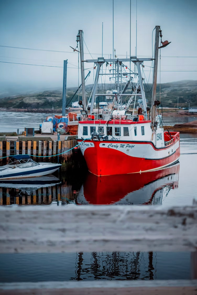 A red-hulled fishing boat lying at a timber wharf in flat calm water, stacked
                  traps and floats on the dock behind her and low hills fading into fog.